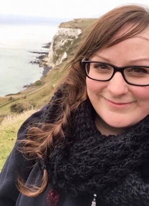 Woman with brown hair in a braid with glasses on the right side of the photo standing in front of green lined cliffs with white rocks looking out to sea at White Cliffs of Dover