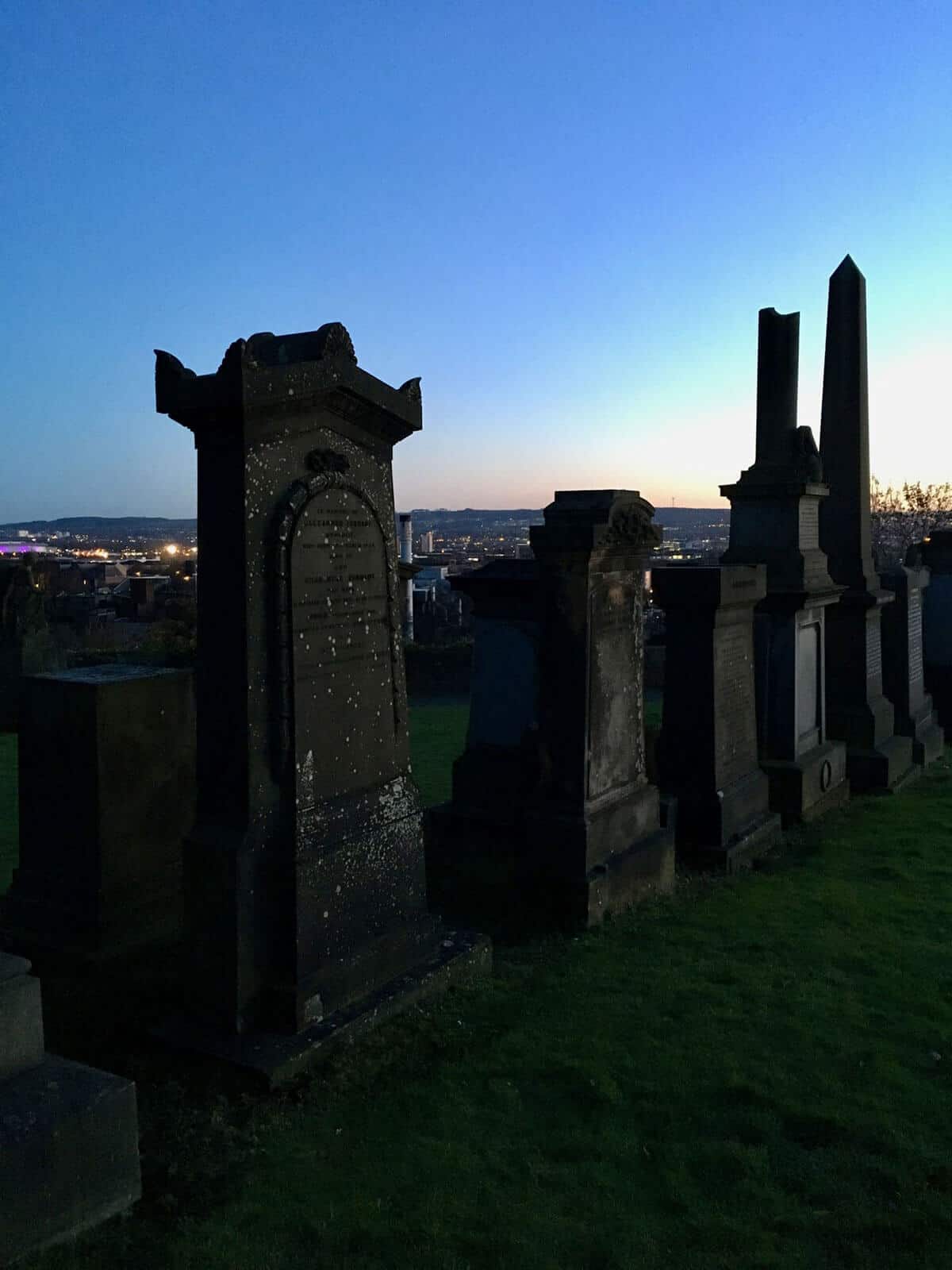 Six aged, mossy and weathered Victorian style grave stones and tombs with the Glasgow skyline in the back ground as the sun sets casting a dark shadow glow on the graves.