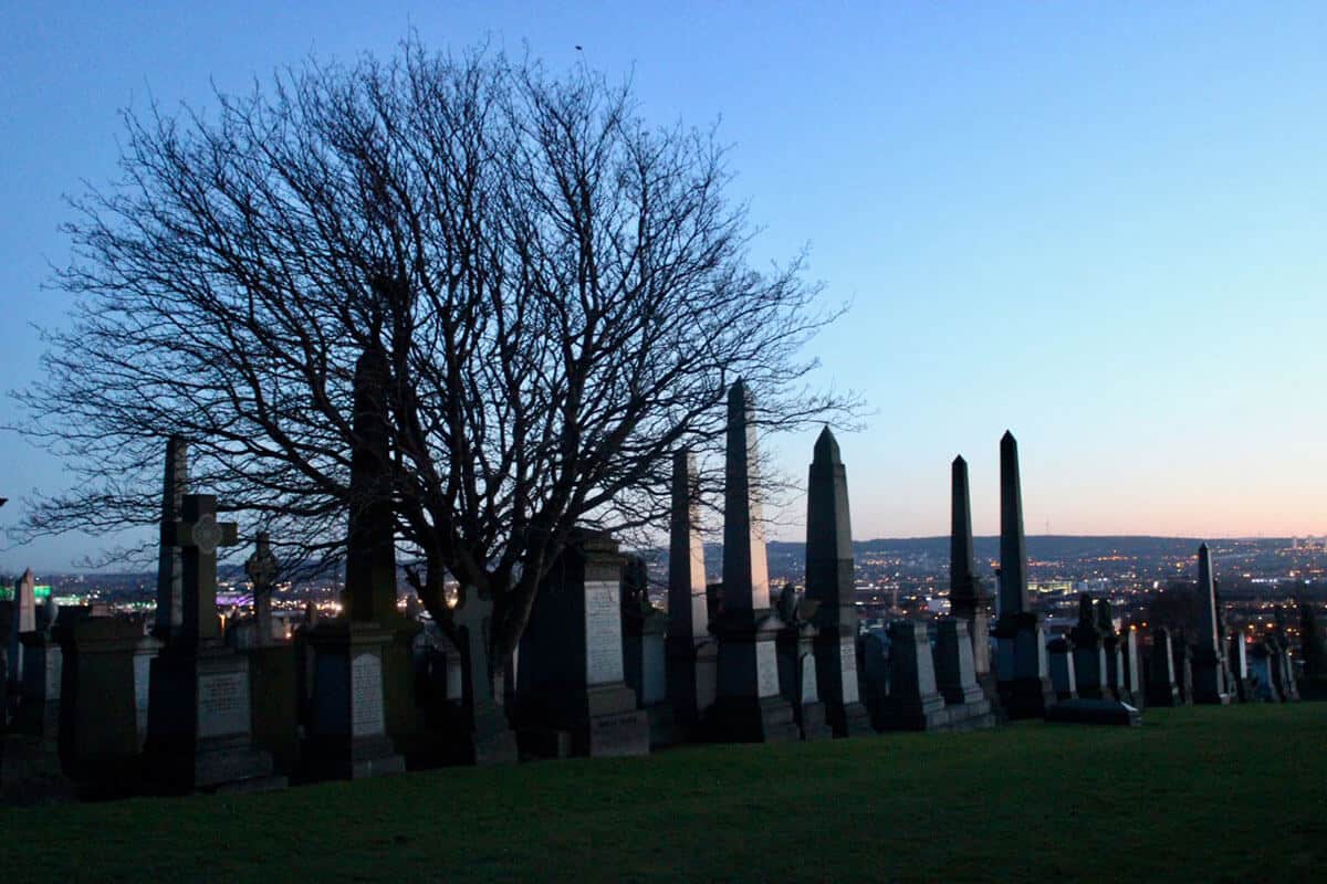 A green hill lined with several graves, cemetery stones and tombstones with a deciduous tree with no leaves sits behind the graves. The sun is setting and a red glow lights up the gravestones in the Glasgow Necropolis