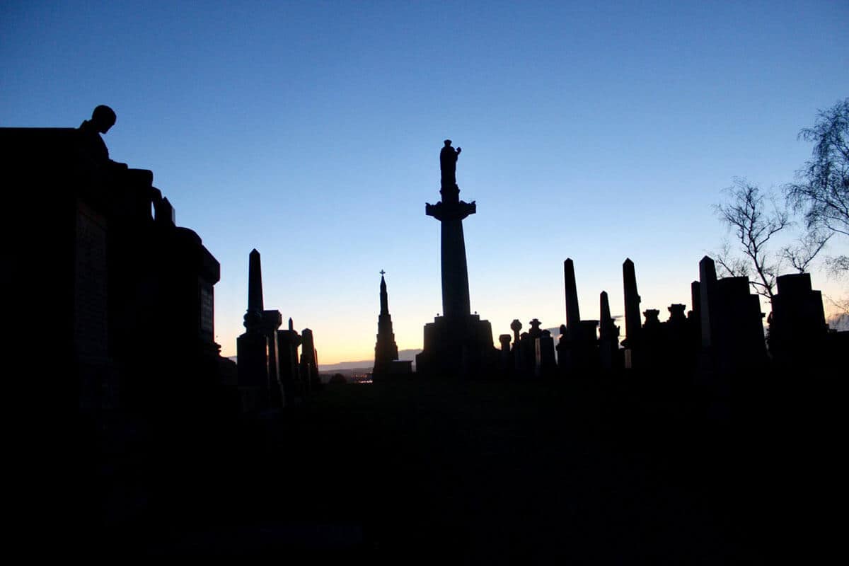 Victorian Graves in black shadows as the sun sets behind with an orange glow and a dark blue sky in the Glasgow Necropolis