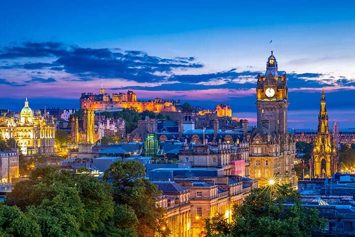 Edinburgh City Scape at dusk, blue and pink sky with buildings lit up. A tower clock on the right side with the Edinburgh Castle in the back. One of the places a foreigner can get a job in Scotland.